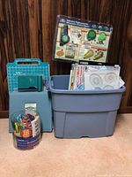 Wide shot showing large plastic bin with assorted crafting materials, paper cutter, storage box, and container of colorful scissors