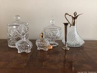 Photo showing all seven crystal items arranged in a row on a wooden surface: a large ice bucket, covered candy dish, two perfume bottles, crystal decanter with silver handle, two small silver-tone vases.