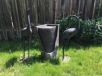 Front view of outdoor metal bird fountain with two crane sculptures placed in grass in backyard near wooden fence.