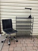 Black office chair and metal shelving unit with adjustable lamp against a siding wall.