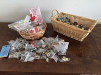 Wide view showing two wicker baskets with assorted pins and many pins in clear plastic bags spread on table.