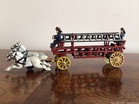 Full side view showing the red fire engine with ladder, two firefighter figures, and two white horses pulling the carriage.