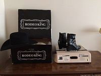Black Rodeo King cowboy hat displayed on top of its branded box alongside a pair of black women's riding boots on the box they came in.