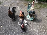 Seven rooster and chicken figurines arranged outdoors on driveway. Large multicolored rooster figurine at back with smaller roosters and chickens grouped around. Shows signs of fading and weather exposure.