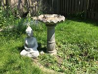 Outdoor photo showing stone/concrete bird bath with scalloped bowl and fluted pedestal alongside a seated Buddha statue on grass near a garden bed with fence in background.
