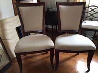 Pair of two beige upholstered dining chairs with dark wood finish shown from front angle on wood floor.