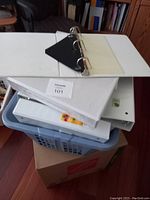 Stack of white binder folders placed in a blue laundry basket on cardboard box. Top folder is open to show three metal rings inside, white exterior, and clear plastic sleeve on the spine.