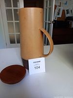 Side view of tall beige and brown pottery pitcher with handle and wooden lid removed, showing lid beside it.