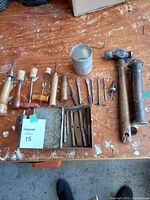 Photo of assorted tinsmithing and metalworking tools laid out on a wooden surface, including hammers, punches, awls, chisels, a metal cup, and a small metal tin.