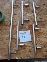 Five silver metal cabinet pulls of varying lengths placed on oriented strand board surface. Each includes threaded screws attached to mounting posts.