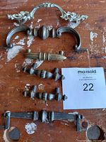 Top-down photo showing six metal antique cabinet pulls in various designs and sizes arranged on a wood surface with MaxSold tag #22.