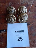 Set of 4 brass-colored antique cabinet knobs with raised flower design placed on a wooden surface and a paper labeled 25.