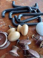 Close-up photo of several black metal pulls, natural wood knobs, reddish brown wooden knobs, and a grey concrete knob on a wooden surface.