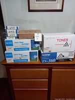 Photo showing multiple toner cartridge boxes including Brother TN-360 box and various blue and white boxes labeled 'High Quality Laser Toner Cartridge' with various TN model numbers.
