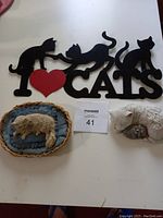 All three cat-themed items shown on white surface: black wall hanging with cats and text, small white curled cat figurine, and light-colored cat figurine on cushions in wicker basket.