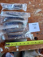 Seven brass wardrobe pull handles in original Eurofit plastic packaging shown on a worn wooden surface. A measuring tape indicates each pull is about 3.5 inches long.