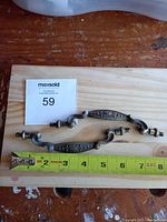Two antique metal cabinet pulls placed on a wooden board with a measuring tape showing the length approximately 6 inches, maxsold lot tag 59 visible.