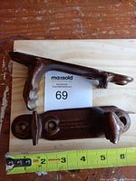 Two antique large metal wall hooks/braces placed side by side on a wooden surface with a measuring tape showing length around 6 inches.