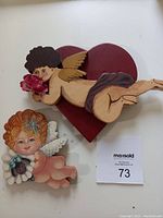 Two wooden cupid wall hangings on white surface: one lying on a red heart, the other holding grapes.