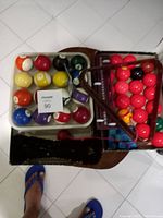 Photo showing various billiard balls in a box, including striped and solid colors, with a carrying case underneath.