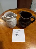 Two artisan pottery pitchers, one white with floral embossing and one black with carved floral design, placed on wooden surface with MaxSold lot number 96 card visible.