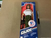 Front view of G.I. Joe Grunt action figure in box showing figure wearing white shirt, green pants, and helmet. Box is blue with various text and promotional sticker.