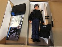 View of complete figure in box with accessories: black vest, communication device, certificate of authenticity, and plastic stand visible