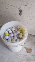 Top view of a large white pail filled with a mix of white and yellow golf balls, mostly used, visible brands include Titleist and Top Flite.
