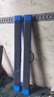 Two folded Mastercraft sawhorses leaning against a wall. Each is blue metal with black rubber top surfaces and black carrying handles on the side.
