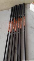 Seven clubs laid side by side showing black grips, orange/black shafts, silver heads