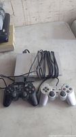 Full view of the Playstation 2 console with two wired controllers (black and silver) connected, cables coiled on top