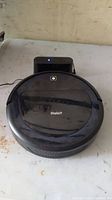Robit robotic vacuum cleaner on a white surface with its charging dock behind it. The vacuum is round, black, with a glossy finish, showing some dust and wear.