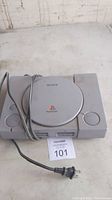 Top view of the grey Sony PlayStation 1 console placed on a white surface with attached power cord and MaxSold lot number 101 tag in front.