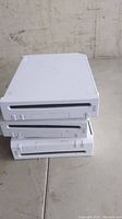 Photo showing three white Nintendo Wii gaming consoles stacked on top of each other on a rough surface.