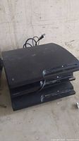 Three black PlayStation 3 consoles stacked on a dusty surface, showing top and front views with power cable visible.