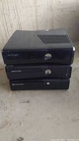 Photo showing three black Xbox 360 consoles stacked on top of each other against a white wall background with visible wear on consoles.