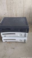 Photo of three Xbox 360 consoles stacked—one black on top and two white underneath, showing general wear and no accessories included.