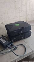 Two original Xbox consoles stacked on a table with one power cable and power brick in front of them.