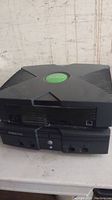 Photo of two stacked original Xbox consoles showing front and top views with visible green logo, black exterior, and multiple front panel ports.
