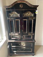 Front view of the black wood armoire showing intricate gold detailing on the doors and top, with two drawers below.