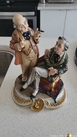 Front angled view of ceramic figurine showing two men, one standing and one seated on an ornate chair, on a decorative base.