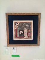 Photo of framed needlepoint wall art showing geometric and floral needlework with cushioned blue mat and gold ornate frame.