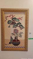 Framed needlepoint floral artwork showing stylized flowers and bonsai-like tree in teal, pink, burgundy, and cream colors with decorative gold frame.