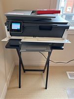 HP OfficeJet Pro 8025 printer on black wood stand with open paper tray and red box on top, next to window with wall outlet visible.