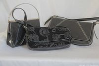 Three black dress purses arranged side by side: Nine West floral purse in front center, KGB black purse with silver handle on left, Ricky's black patent purse with white stitching on right.