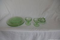 Full lot of clear green uranium or vaseline glass pieces including round plates, pedestal bowls, and an ashtray, displayed on white fabric.