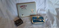 Three tin boxes displayed on white fabric including a Sweet Caporal Cigarettes tin, Barkleys mints tin, and a small R Bell & Co. matchbox tin.