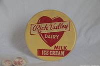 Front view of the large Rich Valley Dairy advertising button in yellow and red, heart logo, with 30 white and red milk bottle caps below.