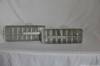 Two vintage metal ice cube trays displayed side by side on white cloth background, showing full trays with segmented compartments.