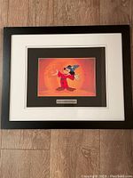 Full view of the framed serigraph cell showing the double matting and black frame on wooden floor.